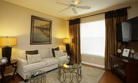 A living room with a white couch, a painting on the wall, a ceiling fan, and a glass coffee table.