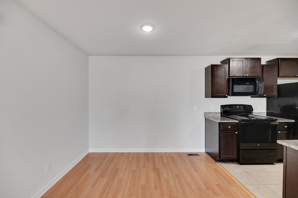 an empty living room with a kitchen with black appliances and wooden floors