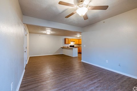an empty living room with a ceiling fan and a kitchen in the background
