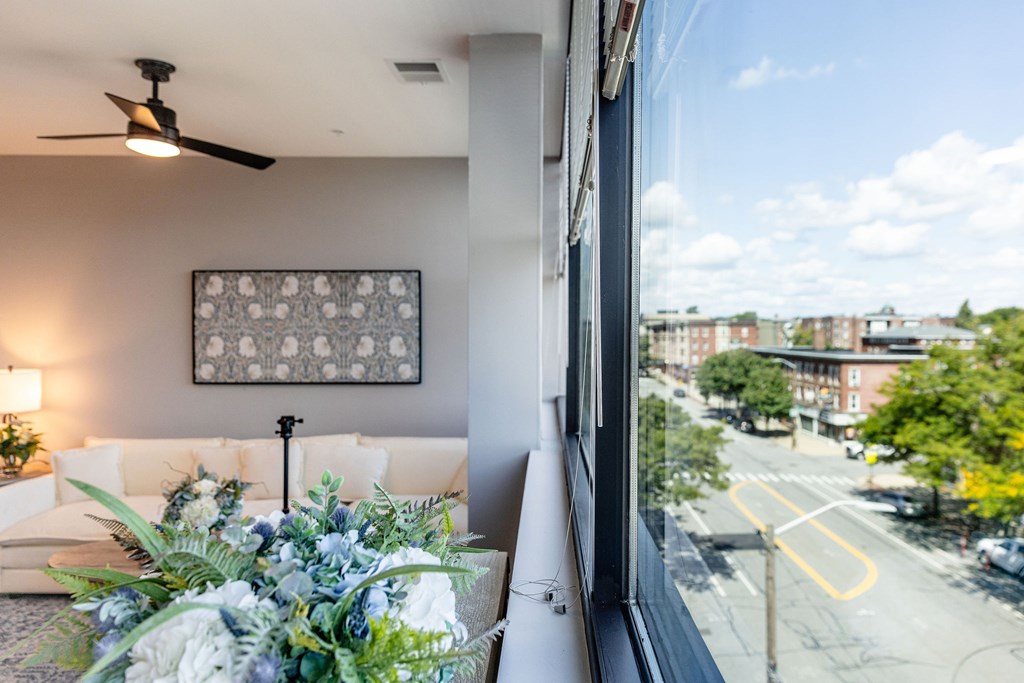 A living room with a couch, a fan, and a window overlooking a street.