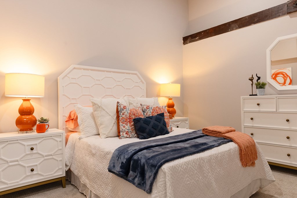 A bedroom with a white bed, a white dresser, and a white nightstand with orange lamps.