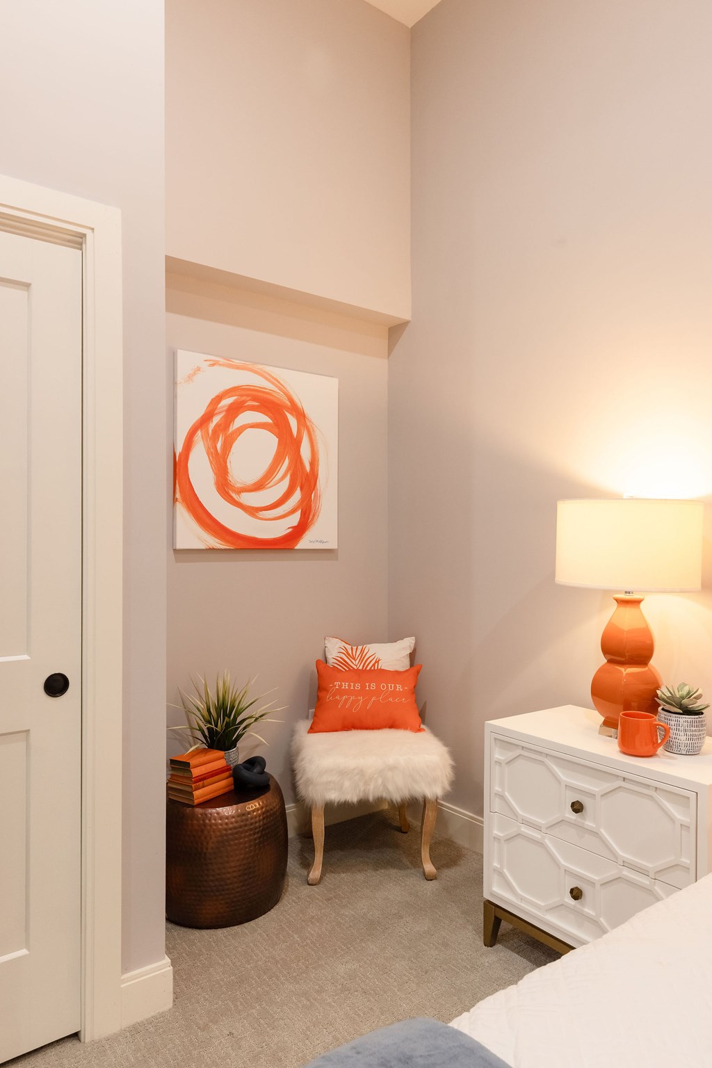 A room with a white chair and a white dresser with an orange lamp and orange pillow.