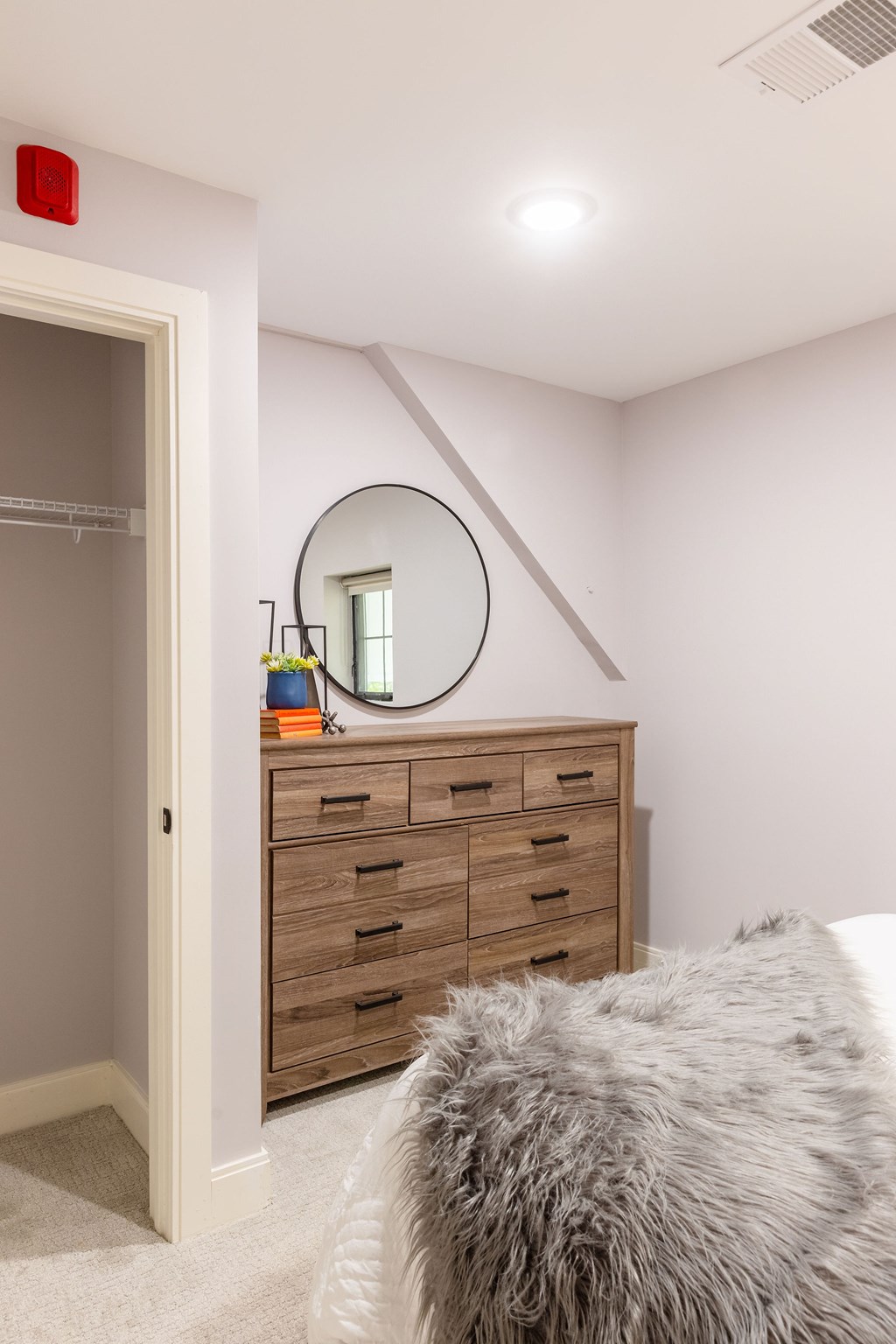 A bedroom with a grey furry bedspread and a wooden dresser with a round mirror above it.
