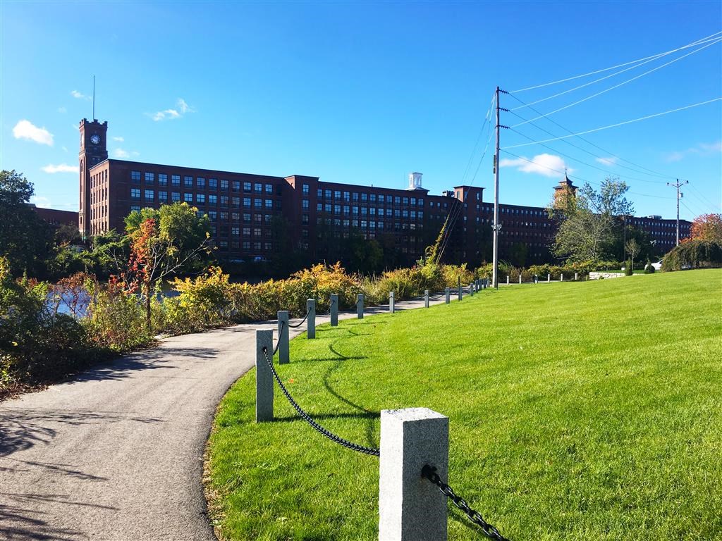A large building with a clock tower is behind a grassy area with a walking path.
