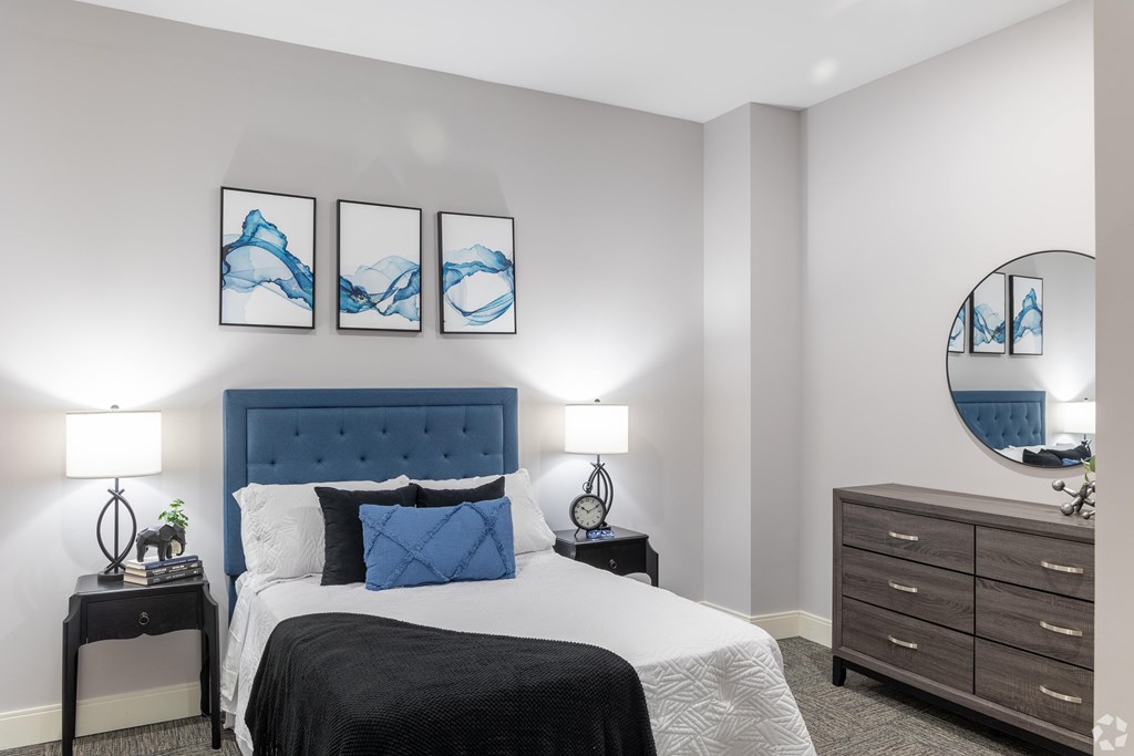 A bedroom with a large bed, a nightstand with a lamp, and a dresser with a mirror.