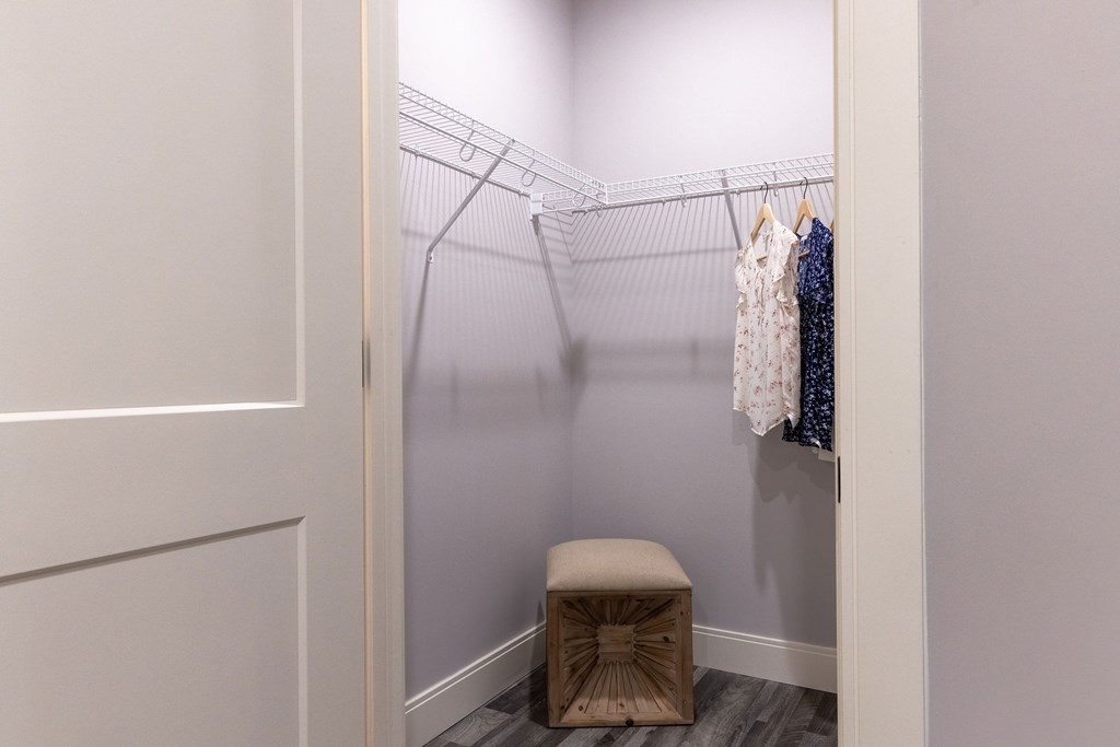 A small closet with clothes hanging on a wire rack and a stool in front.