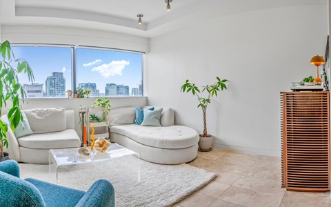 A living room with a white couch, a glass table, and a large window with a view of the city.