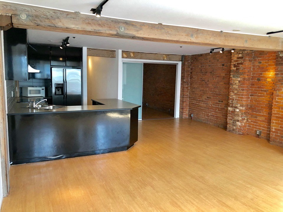 an empty living room with a kitchen and a exposed brick wall