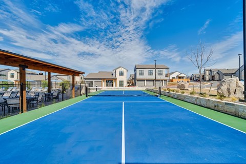 A tennis court with a blue surface and white lines is surrounded by houses and trees.