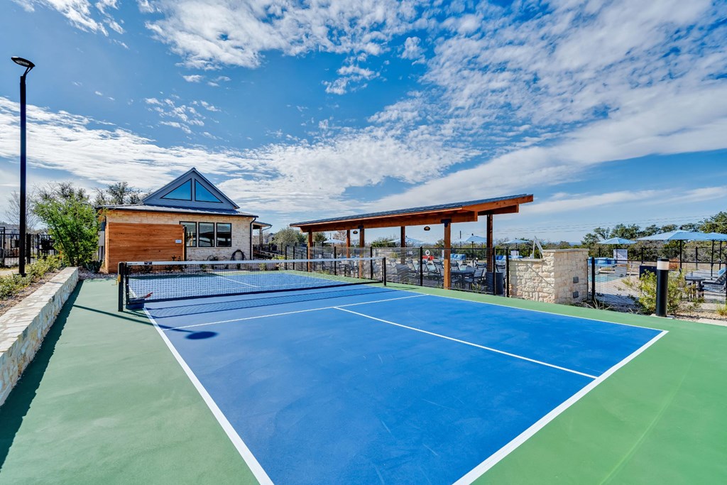 A tennis court with a blue surface and white lines, surrounded by a fence and a building with a blue roof.