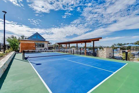 A tennis court with a blue surface and white lines, surrounded by a fence and a building with a blue roof.