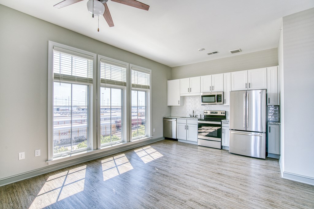 an empty living room with a large window and a kitchen with stainless steel appliances
