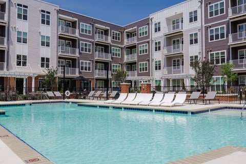 A swimming pool is surrounded by lounge chairs in front of apartment buildings.