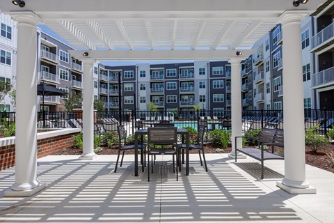 A white pergola with a table and chairs is in front of apartment buildings.