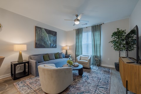 A living room with a grey couch, a green lamp, a painting on the wall, a ceiling fan, a window with curtains, a small table with a vase of flowers, a chair, and a television.