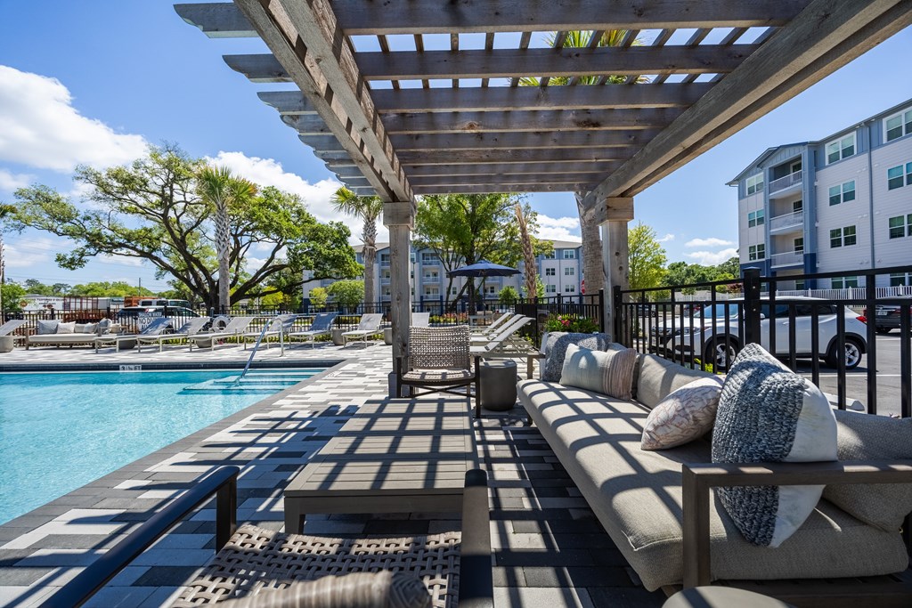 A poolside area with a wooden pergola and a row of cushioned seating.