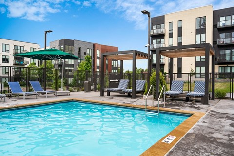 A swimming pool surrounded by chairs and a fence in front of apartment buildings.