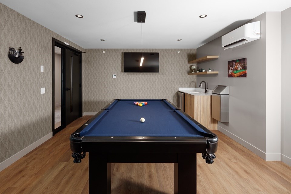 A pool table is in the middle of a room with a television and a bar in the background at The Ambrosi in Kelowna, BC.