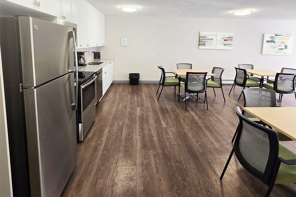 Social room with couch, tables, chairs, and stainless steel appliances at Brockville Apartments in Brockville, ON