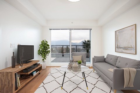 A living room with a grey couch, a wooden entertainment center, and a large wcape at 388 Kaslo East Village Apartmentindow overlooking mountain landss.