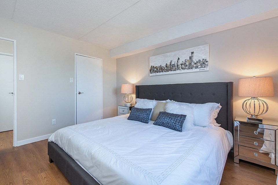 Primary bedroom, looking towards closet, with a grey upholstered bed frame and white linens at Lorneville Apartments in Cornwall, ON