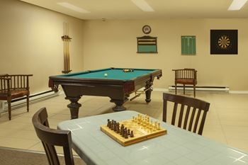 Games room, showing pool / billiard's table, with a dart board in the background and a table with a chess set in the foreground at Lorneville Apartments in Cornwall, ON