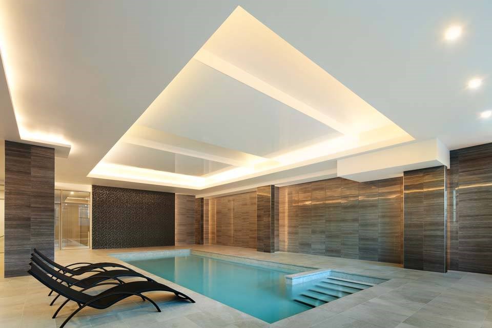A modern indoor swimming pool with a wooden wall and black lounge chairs.