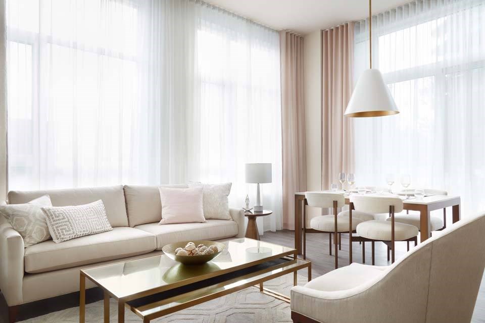 A modern living room with a white sofa, a glass coffee table, and a dining area in the background.