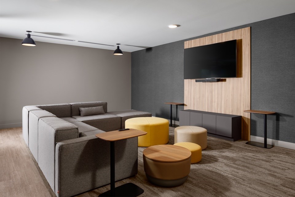 A modern social room with a grey sofa, yellow ottoman, and a wooden tv stand at The Ambrosi in Kelowna, BC.