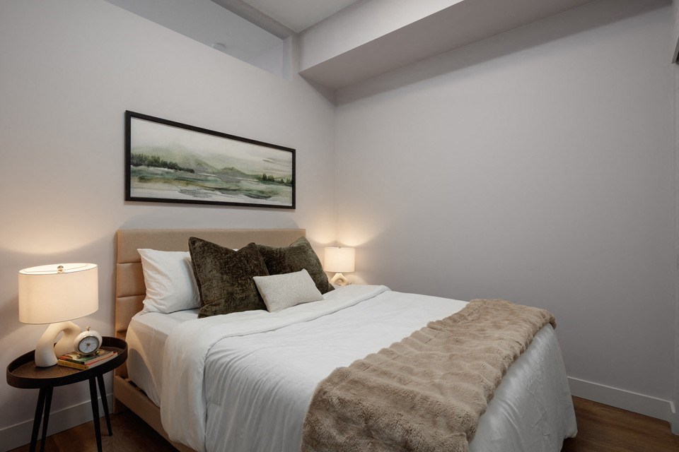A bedroom with a bed, pillows, a blanket, a nightstand with a lamp, and a painting on the wall at The Ambrosi in Kelowna, BC.