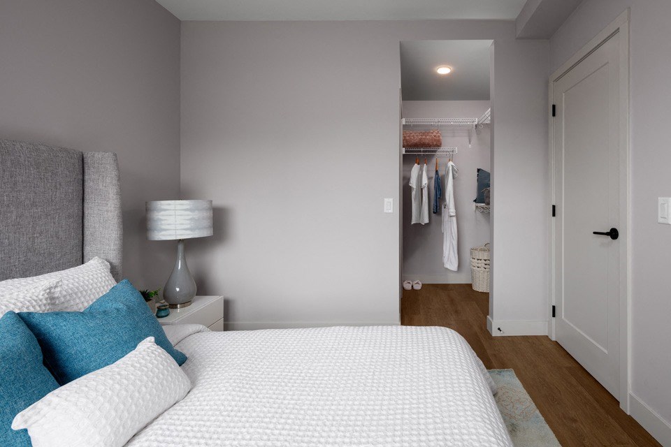 A bedroom with a bed, pillows, a lamp, and a closet at The Ambrosi in Kelowna, BC.