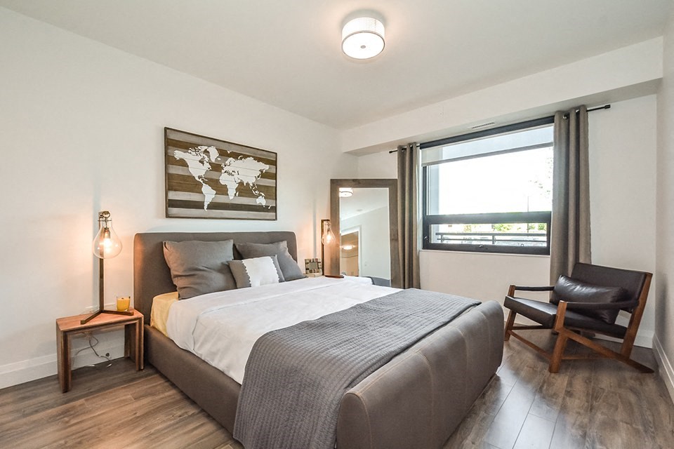 Bedroom with a large, grey upholstered bed and a window in a 1 bed 1 bath variation B in building A at Trio on Belmont in Kitchener, ON