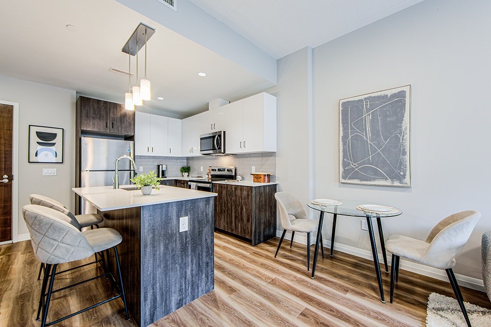 Dining area and kitchen with a two-seater dining table, kitchen island, wood-look lower, white upper and stainless steel appliances in a 2 bedroom 2 bathroom variation C layout at Vale Station Apartments in Waterloo, Ontario