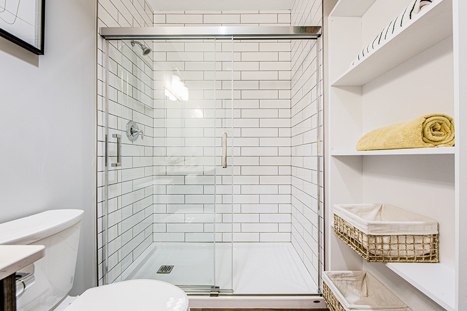 Main bathroom with a white tiled shower with a glass door, white toilet and shelving in a 2 bedroom 2 bathroom variation C layout at Vale Station Apartments in Waterloo, Ontario.