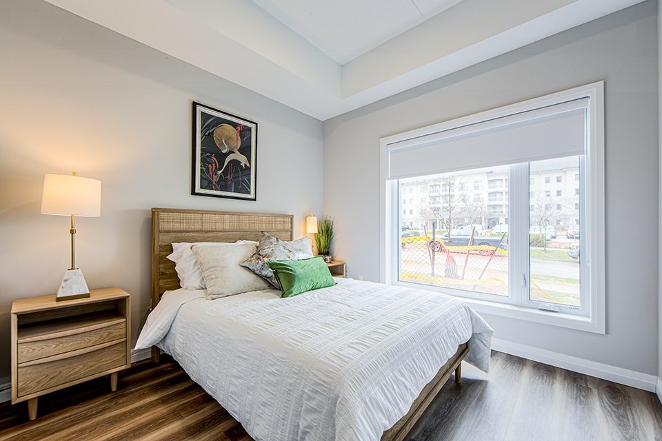 Second bedroom with a large bed, a nightstand with a lamp, and a picture on the wall next to a large widow with blinds in a 2 bedroom 2 bathroom variation C layout at Vale Station Apartments in Waterloo, Ontario