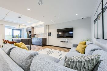 Large grey sectional couch and flat screen TV with a kitchen in the background in the social room at Vale Station Apartments in Waterloo, Ontario