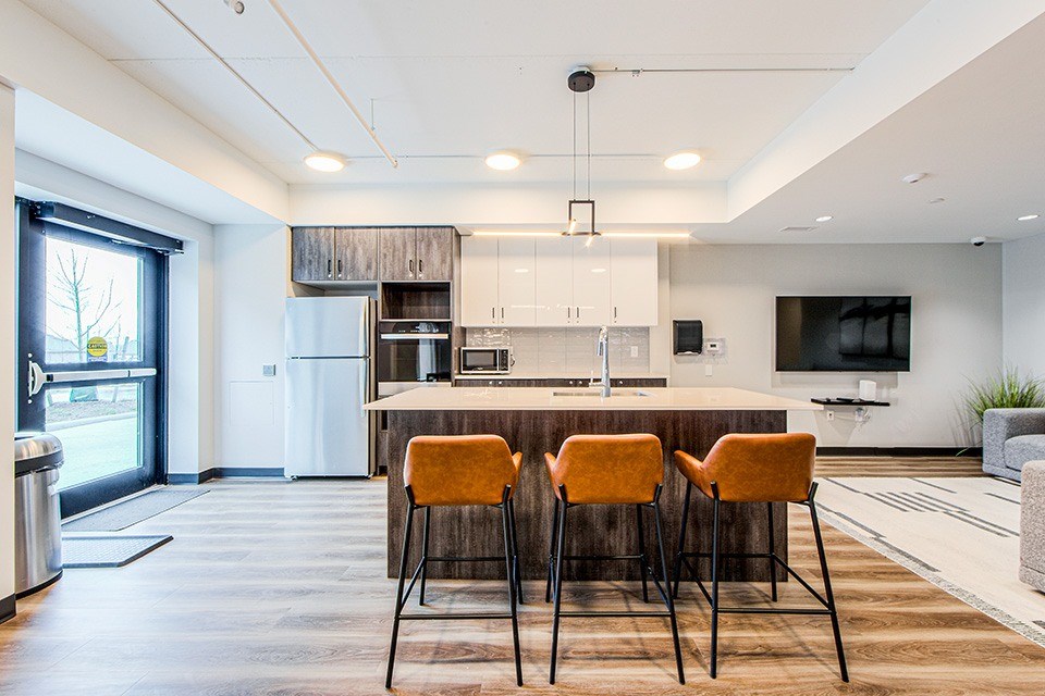 Kitchen located in the social room with wood-look lower and white upper cabinets, a large kitchen island with three bar stools and stainless steel kitchen appliances at Vale Station Apartments in Waterloo, Ontario