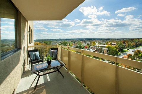 Edgehill Apartments in Barrie, ON balcony view overlooking lush trees and homes. The weather is clear with bright blue skies and two chairs are sat next to each other with a glass table in front.
