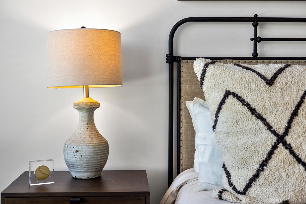 A lamp with a white shade sits on a nightstand next to a bed with a white and black patterned pillow.