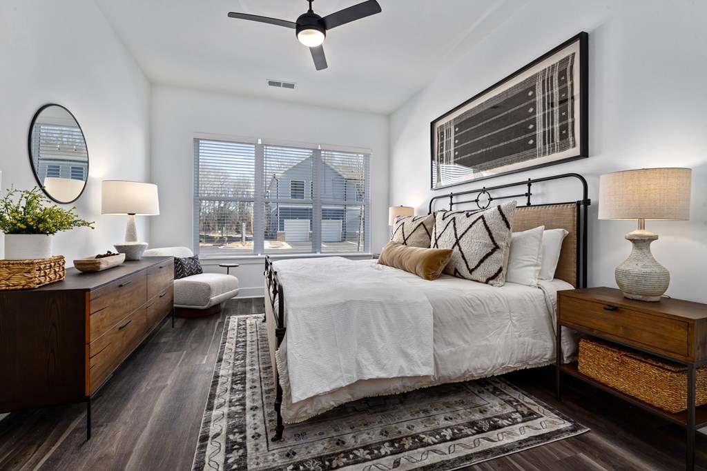 A bedroom with a large bed, a ceiling fan, and a window with a view of a house.