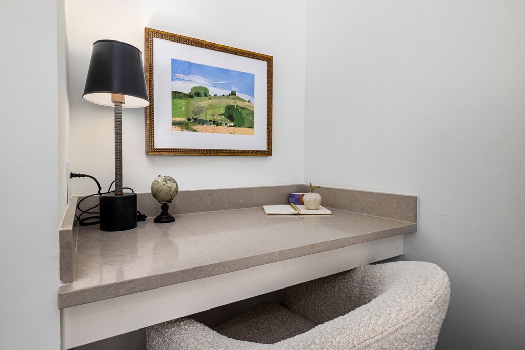 A picture of a landscape is framed and hung on a wall above a desk with a lamp and other items on it.