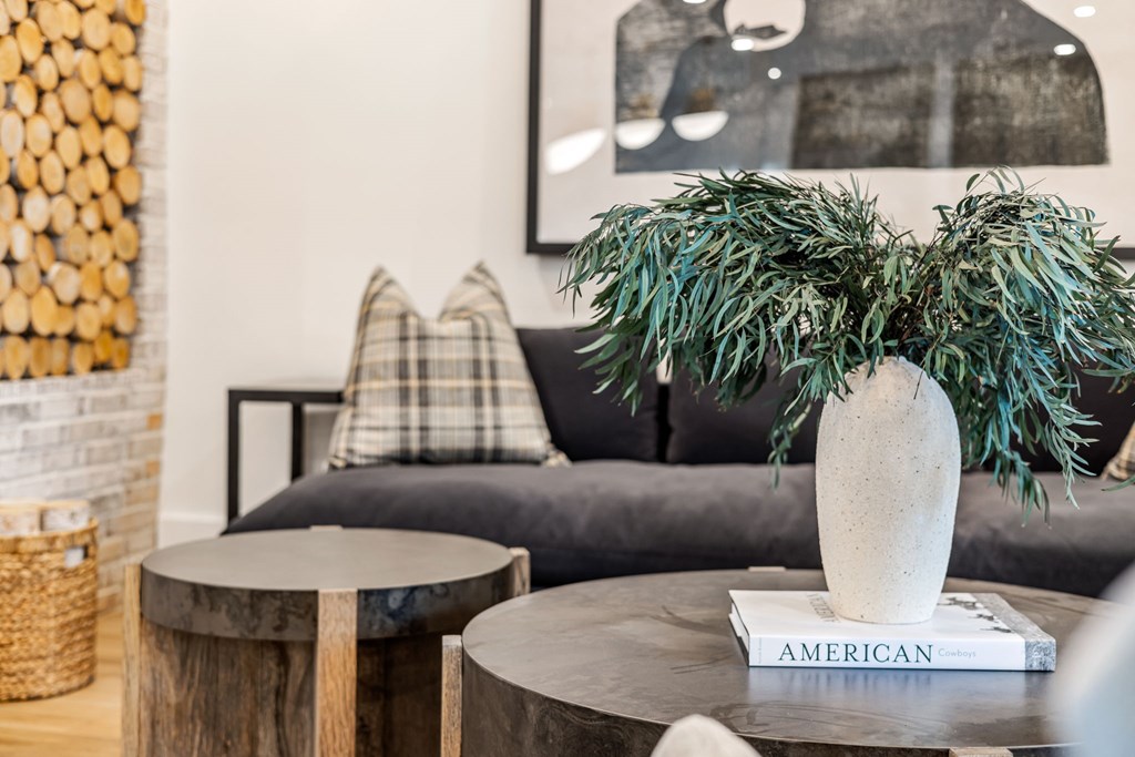 A living room with a grey couch, a wooden coffee table, and a vase with green plants on top of a book titled "American".