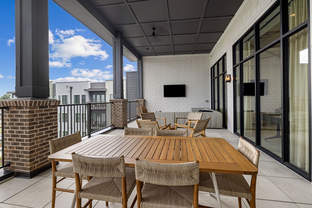 A wooden table with chairs is on a patio with a brick pillar and a glass door.