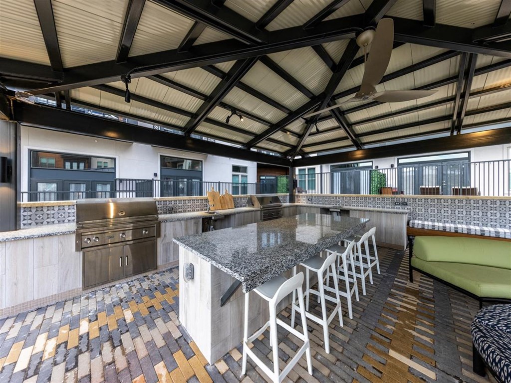 An outdoor kitchen under a covered patio with two barbecue grills, a sink, granite countertops, a ceiling fan, and an island with barstools near a lounge area.