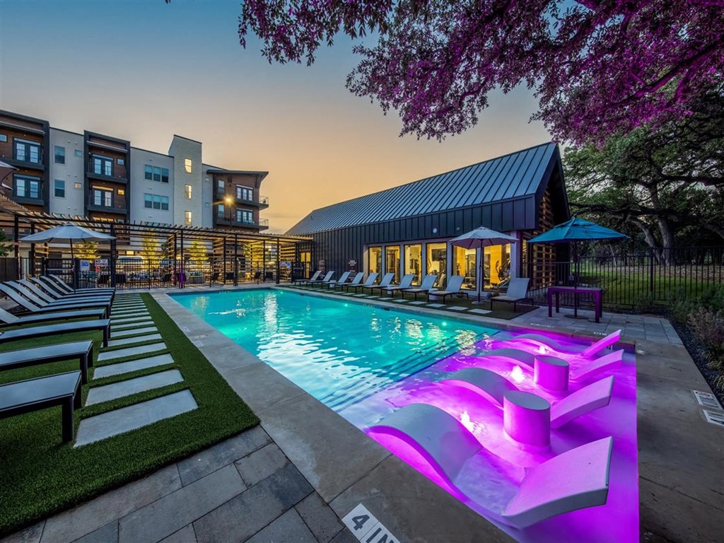 A resort-style pool surrounded by lounge chairs and umbrellas near a community gym and apartment buildings.
