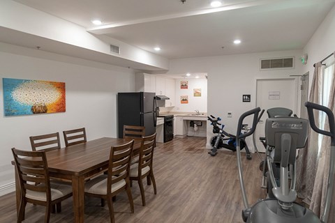 A room with a dining table, chairs, a refrigerator, a kitchenette, and a treadmill.
