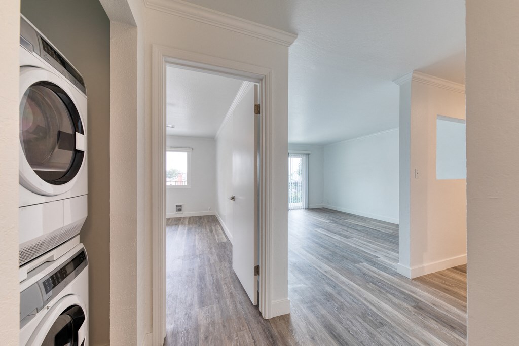 Apartment interior door open leading to bedroom and living room open floorplan. Stackable white washer and dryer on the side of the hallway.
