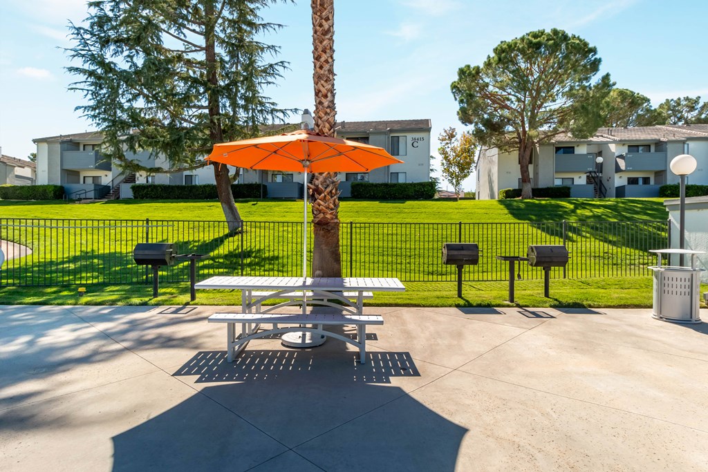 A picnic table with an orange umbrella is in the middle of a concrete area with a tree and mailboxes in the background at Chaparral Apartments, Palmdale, CA, 93551