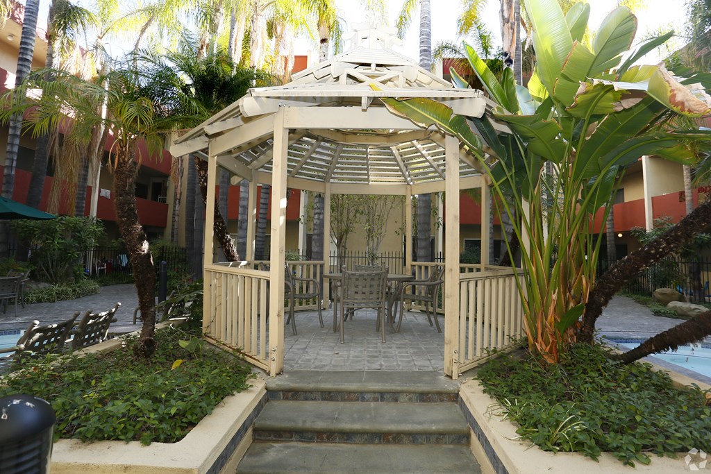 A gazebo with a table and chairs is surrounded by plants.