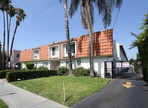 A white house with a red tile roof is surrounded by greenery and palm trees.
